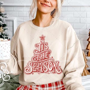 Op de afbeelding: Beige sweatshirt met een rood kerstboomontwerp en de tekst "Tis the Season". De trui wordt gecombineerd met een rood-zwarte geruite broek. Een witte mok met rode stippen wordt vastgehouden. Kerstboomversieringen op de achtergrond.