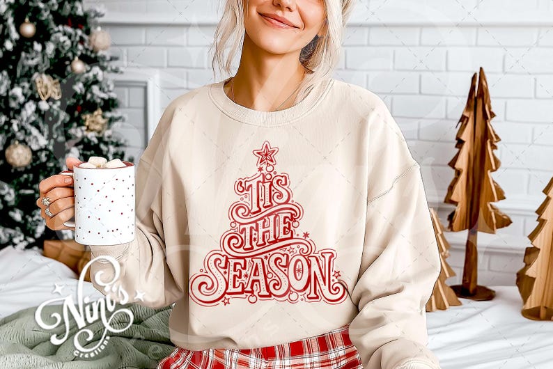 Op de afbeelding: Beige sweatshirt met een rood kerstboomontwerp en de tekst "Tis the Season". De trui wordt gecombineerd met een rood-zwarte geruite broek. Een witte mok met rode stippen wordt vastgehouden. Kerstboomversieringen op de achtergrond.