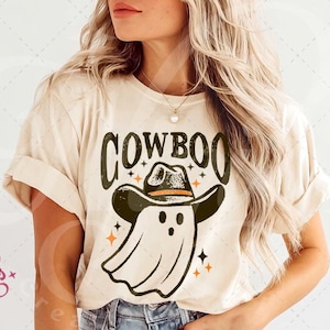 Puede incluir: Camiseta beige con la palabra "COWBOO" en letras verde oscuro. El dise&ntilde;o presenta un fantasma con sombrero de vaquero y estrellas naranjas. Una camiseta gr&aacute;fica con tem&aacute;tica de Halloween.