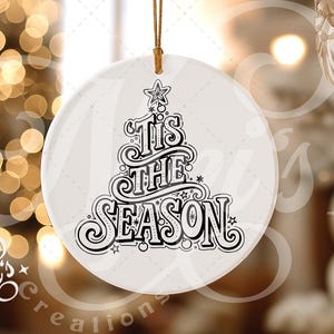 Op de afbeelding: Een witte, ronde kerstversiering met de tekst "'Tis the Season" in zwarte letters. Het ontwerp bevat een kerstboom en sterren. Een gouden draad is bevestigd om op te hangen. De achtergrond toont vage kerstverlichting.