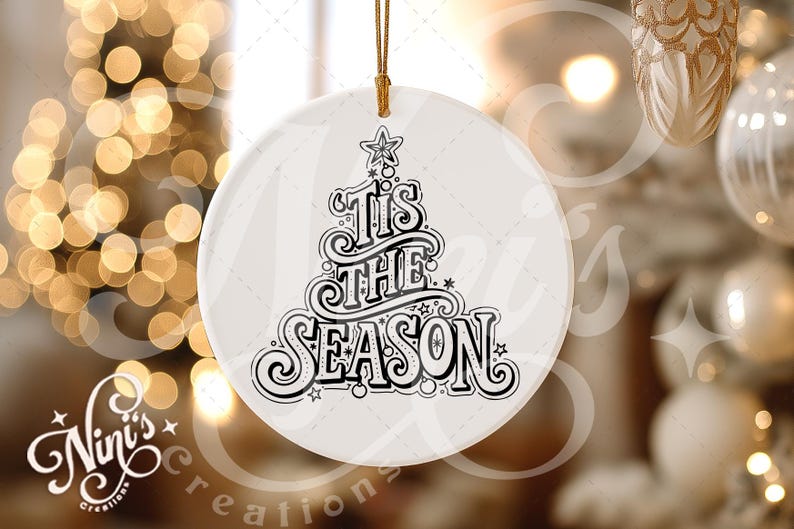 Op de afbeelding: Een witte, ronde kerstversiering met de tekst "'Tis the Season" in zwarte letters. Het ontwerp bevat een kerstboom en sterren. Een gouden draad is bevestigd om op te hangen. De achtergrond toont vage kerstverlichting.