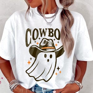 Puede incluir: Camiseta blanca con un gr&aacute;fico que presenta la palabra "COWBO" sobre un fantasma con sombrero de vaquero. El dise&ntilde;o incluye estrellas naranjas. La camiseta es de estilo casual y unisex.