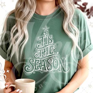 Op de afbeelding: Een groen t-shirt met de tekst "Tis the Season" in een kerstboomontwerp. Het ontwerp is wit en bevat sterren en krullen. Het shirt is effen van kleur en heeft opgerolde mouwen.