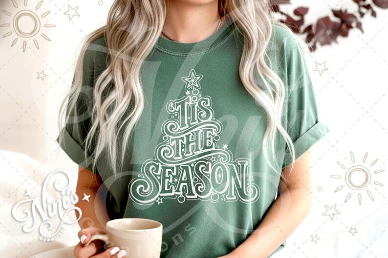 Op de afbeelding: Een groen t-shirt met de tekst "Tis the Season" in een kerstboomontwerp. Het ontwerp is wit en bevat sterren en krullen. Het shirt is effen van kleur en heeft opgerolde mouwen.