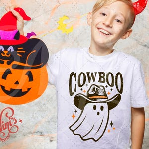 Puede incluir: Un ni&ntilde;o lleva una camiseta blanca con la palabra "COWBOO" y un dibujo de un fantasma con sombrero de vaquero. El ni&ntilde;o sostiene una decoraci&oacute;n de Halloween de un gato negro en una calabaza con sombrero de bruja.