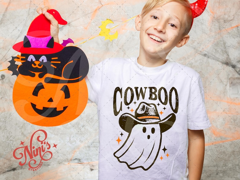 Puede incluir: Un ni&ntilde;o lleva una camiseta blanca con la palabra "COWBOO" y un dibujo de un fantasma con sombrero de vaquero. El ni&ntilde;o sostiene una decoraci&oacute;n de Halloween de un gato negro en una calabaza con sombrero de bruja.