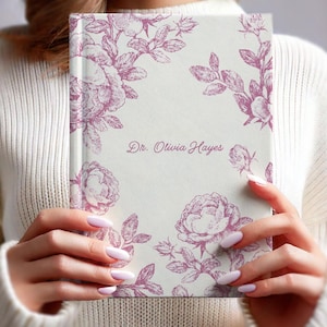 May include: A hardcover journal with a cream-colored cover featuring a floral design in shades of purple. The text "Dr. Olivia Hayes" is printed on the cover in a matching purple script. The journal is held by a person wearing a white sweater.