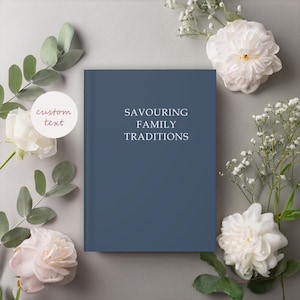 K&ouml;nnte beinhalten: Ein dunkelblaues Hardcover-Journal mit dem Text "Savouring Family Traditions" auf dem Einband. Das Journal ist von wei&szlig;en Blumen und gr&uuml;nen Bl&auml;ttern umgeben.