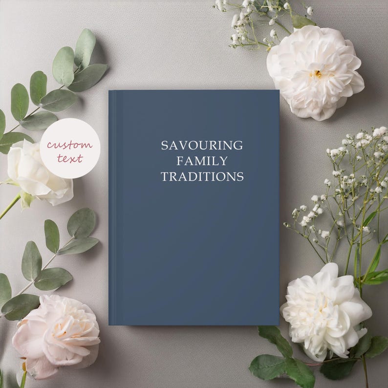 K&ouml;nnte beinhalten: Ein dunkelblaues Hardcover-Journal mit dem Text "Savouring Family Traditions" auf dem Einband. Das Journal ist von wei&szlig;en Blumen und gr&uuml;nen Bl&auml;ttern umgeben.