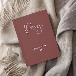 K&ouml;nnte beinhalten: Ein burgunderfarbenes Gebetsbuch mit dem Wort "Pray" in wei&szlig;er Kursivschrift. Das Buch enth&auml;lt au&szlig;erdem den Text "Sophia's Prayers" und ein kleines wei&szlig;es Herz.