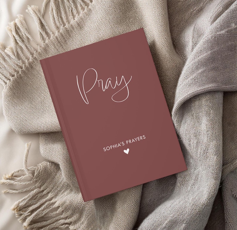 K&ouml;nnte beinhalten: Ein burgunderfarbenes Gebetsbuch mit dem Wort "Pray" in wei&szlig;er Kursivschrift. Das Buch enth&auml;lt au&szlig;erdem den Text "Sophia's Prayers" und ein kleines wei&szlig;es Herz.