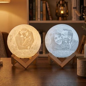 May include: Two spherical moon lamps resting on wooden stands. The left lamp emits a warm yellow light and features a 3D-printed image of a couple embracing. The right lamp emits a cool white light and displays a 3D-printed image of a couple kissing.