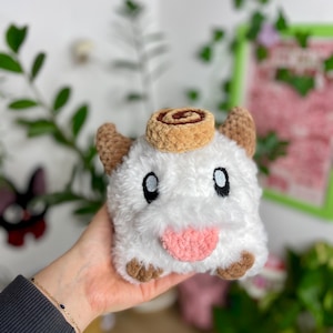 May include: A plush, white creature with brown ears, feet, and a cinnamon roll on its head. The creature has large black eyes and a pink tongue sticking out. The plush toy is held in a hand.