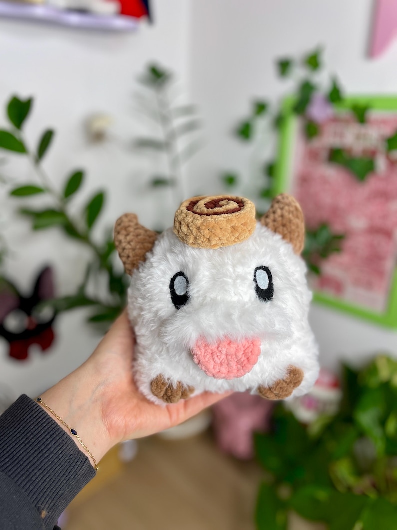 May include: A plush, white creature with brown ears, feet, and a cinnamon roll on its head. The creature has large black eyes and a pink tongue sticking out. The plush toy is held in a hand.