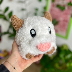 May include: A handmade plush toy in the shape of a Poro, a creature from the game League of Legends. The toy is white with brown ears and feet, black eyes, and a pink tongue. The plush is held in a hand.