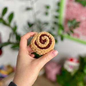 May include: A handmade, plush cinnamon roll in shades of tan and brown. The soft, textured roll is held in a hand, showcasing the detailed spiral design. The background is blurred, with hints of green foliage and a white wall.
