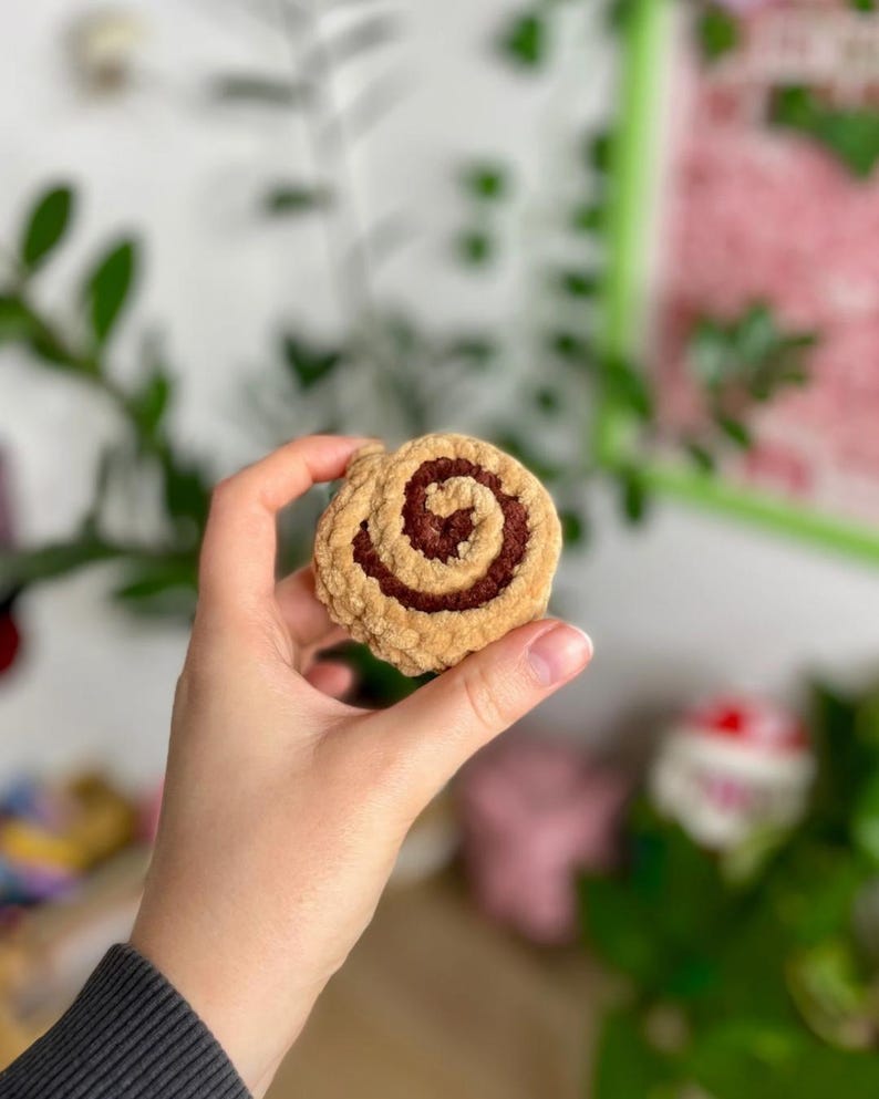 May include: A handmade, plush cinnamon roll in shades of tan and brown. The soft, textured roll is held in a hand, showcasing the detailed spiral design. The background is blurred, with hints of green foliage and a white wall.