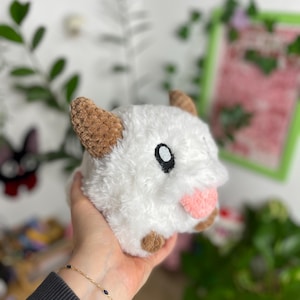 May include: A plush, white stuffed animal with brown ears and feet, and a pink tongue. The toy has a black eye and is held in a person's hand. The background includes greenery and a framed picture.