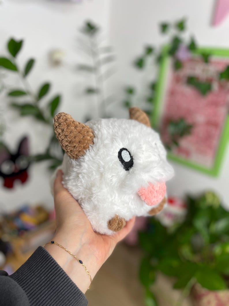 May include: A plush, white stuffed animal with brown ears and feet, and a pink tongue. The toy has a black eye and is held in a person's hand. The background includes greenery and a framed picture.
