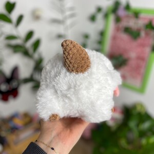 May include: A fluffy white plush toy shaped like a sheep, with brown ears and hooves. The toy is held in a hand, with a gold bracelet visible. The background is a soft focus of greenery and a white wall.