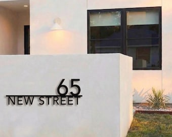 May include: Black metal house number sign with the number 65 and the words "NEW STREET" below. The sign is mounted on a white stucco wall. The background includes a modern house with a black window frame and a small patch of grass.