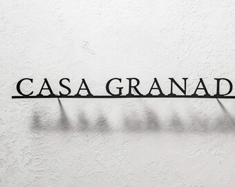 May include: Black metal sign that reads "CASA GRANADA" in a serif font. The sign is mounted on a textured white wall, casting a shadow below the letters. The sign is a decorative element.