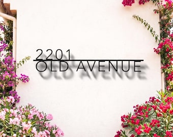 May include: A black metal house address sign mounted on a light-colored wall, displaying the text 2201 OLD AVENUE in a clean, sans-serif font. The sign is positioned above a terracotta pot filled with vibrant red flowers and green foliage.