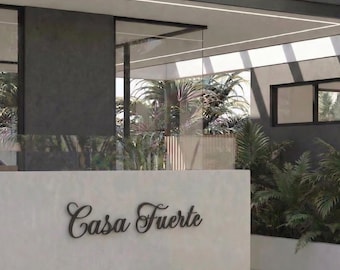 May include: Exterior view of a modern building with a white wall featuring the text "Casa Fuerte" in elegant script. The building has large glass windows and is surrounded by lush greenery, creating a contemporary architectural design.