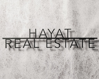 May include: Black metal sign with the words "HAYAT REAL ESTATE" in a modern, minimalist font. The sign is mounted on a textured, light gray background, creating a contrast between the dark text and the lighter surface.