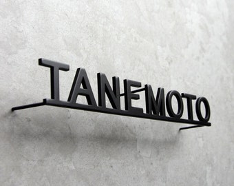 May include: A black metal wall sign featuring the word TANEMOTO in a bold, sans-serif font. The letters are connected by a thin horizontal bar at the base, which extends slightly to the sides to mount the sign away from the wall surface.