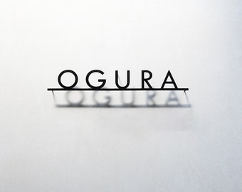 May include: A black metal wall sign featuring the word OGURA in a clean, sans-serif font. The letters are connected by a horizontal bar at the base, casting a soft, blurred shadow of the text directly beneath it on the light-colored wall surface.