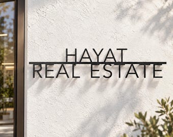 May include: A black metal wall sign featuring the text HAYAT REAL ESTATE in a clean, sans-serif font. The letters are mounted on a horizontal bar and attached to a textured white wall, casting distinct shadows in the sunlight.