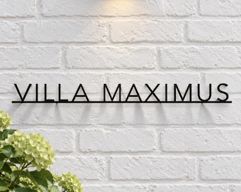 May include: A black metal wall sign featuring the text VILLA MAXIMUS in a clean, sans-serif font. The letters are connected by a thin horizontal bar at the base, creating a linear design mounted against a white brick wall surface.