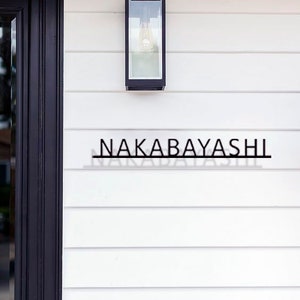 May include: A black metal house number sign with the name "NAKABAYASHI" on a white clapboard siding with a black light fixture above.