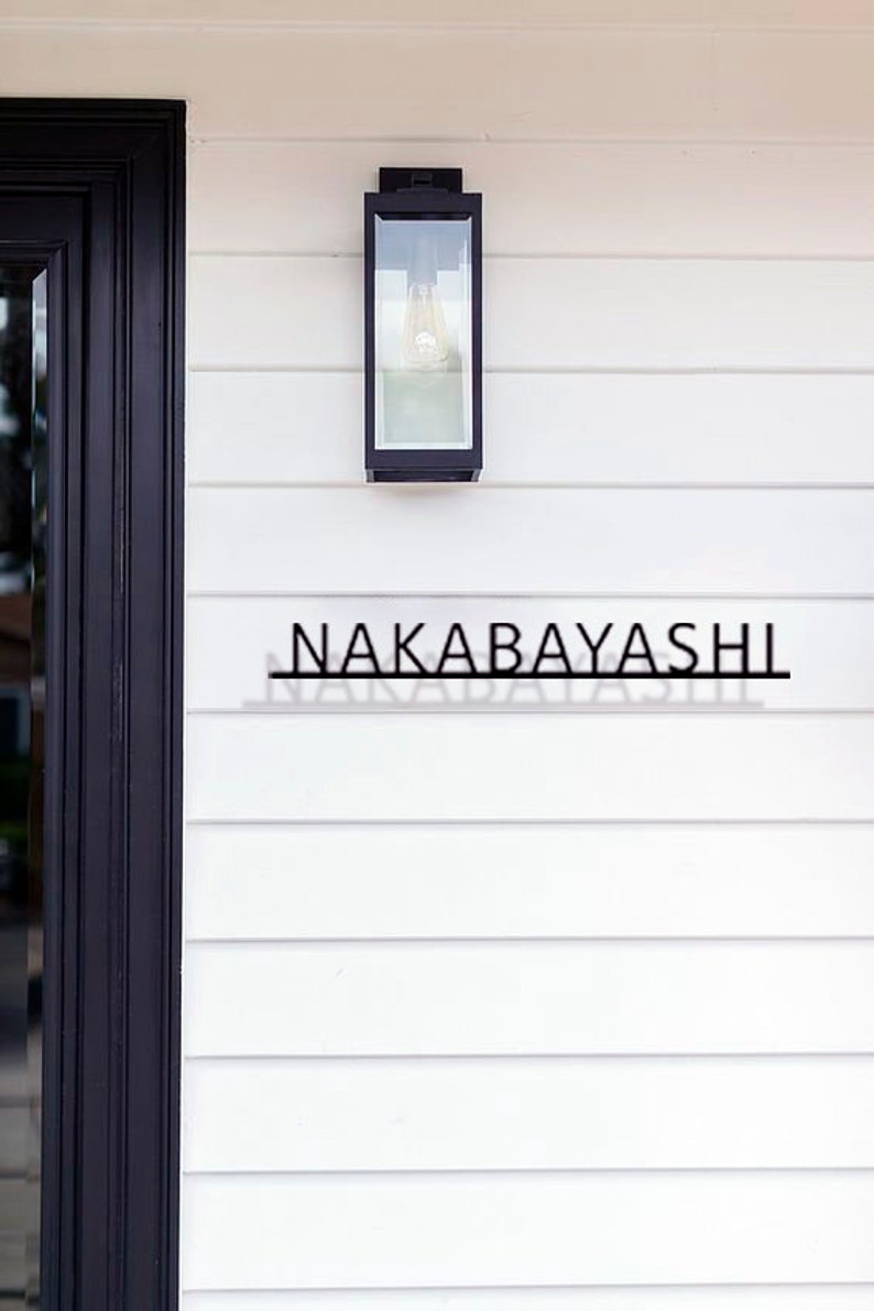 May include: A black metal house number sign with the name "NAKABAYASHI" on a white clapboard siding with a black light fixture above.