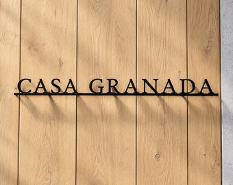 May include: A black metal wall sign featuring the text CASA GRANADA in a bold, serif font. The letters are connected by a thin horizontal line at the base, creating a structured, linear appearance that stands out against the light wood background.