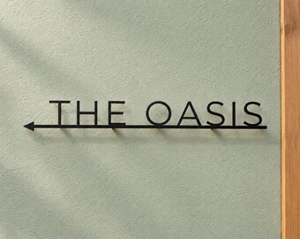 May include: A black metal wall sign featuring the text THE OASIS in capital letters, underlined by a horizontal line that terminates in a left-pointing arrow on the left side, mounted against a textured light green wall surface.