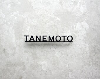 May include: Black metal sign with the name "TANEMOTO" in a sans-serif font, mounted on a light gray, textured wall. The letters are connected by a thin horizontal line.