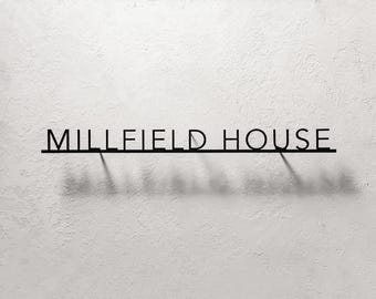 May include: A black metal wall sign featuring the text MILLFIELD HOUSE in a clean, sans-serif font. The letters are connected by a thin horizontal bar at the base, and the design casts a distinct shadow on the surface behind it.