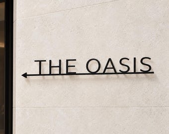May include: A black metal wall sign featuring the text THE OASIS in capital letters. A horizontal line runs beneath the text, extending to the left and ending in a directional arrow pointing left, mounted on a light beige stone wall.