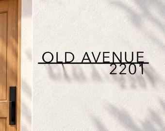 May include: A black metal wall sign featuring the text OLD AVENUE positioned above the number 2201. The letters and numbers are connected by a horizontal line, creating a clean, modern aesthetic mounted on a textured white wall.