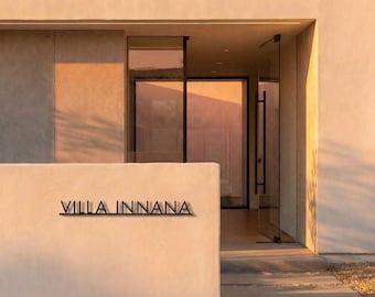 May include: Exterior view of a modern building with a minimalist design. The entrance features glass doors and windows, with the name "VILLA INNANA" in black lettering on a white wall. The building is painted in neutral tones.