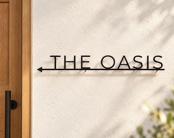 May include: A black metal wall sign featuring the text THE OASIS in capital letters, underlined by a horizontal line that terminates in a left-pointing arrow. The sign is mounted on a textured off-white wall surface.
