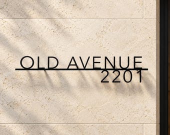 May include: A black metal house sign featuring the text OLD AVENUE in capital letters, with the number 2201 positioned below the text on the right side, all mounted on a light beige stone wall with a horizontal line accent.