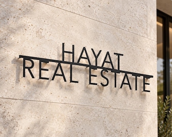 May include: A black metal sign mounted on a beige stone wall featuring the text HAYAT REAL ESTATE in a bold, sans-serif font. The sign consists of two horizontal lines of lettering with a thin black bar connecting the words in a clean layout.