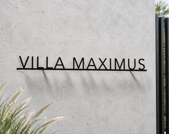 May include: A black metal wall sign featuring the text VILLA MAXIMUS in a clean, sans-serif font. The letters are connected by a thin horizontal line at the base, creating a singular piece of signage mounted against a light gray concrete wall.