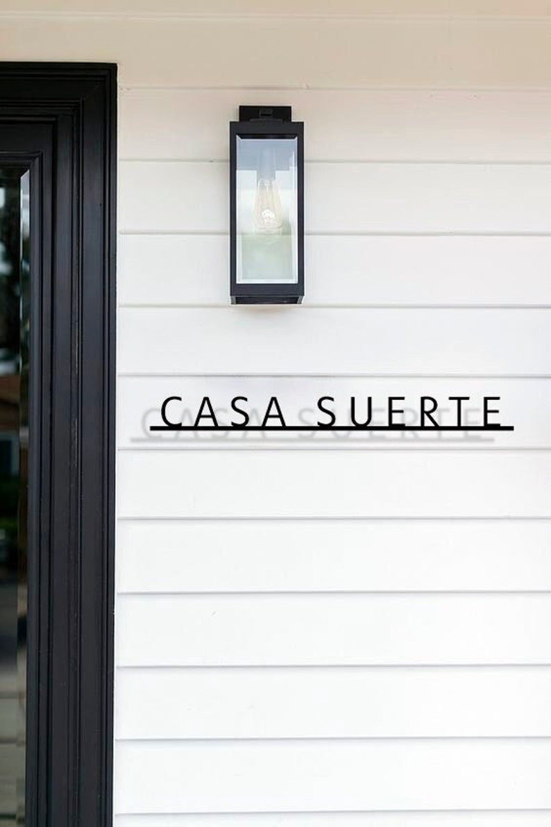 May include: A black metal light fixture with a glass shade is mounted on a white clapboard wall. The text "CASA SUERTE" is displayed on the wall below the light fixture.