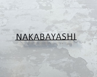 May include: A black metal sign with the name "NAKABAYASHI" in a sans-serif font. The sign is mounted on a textured, off-white wall with visible imperfections and peeling paint, creating a rustic aesthetic.