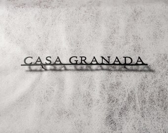 May include: Black metal sign that reads "CASA GRANADA" in a stylized font. The sign is mounted on a textured, off-white surface. The letters are connected by a thin bar.