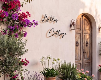 May include: A black metal wall sign with the script text Bellas Cottage mounted on a light-colored stucco wall. The scene includes a wooden arched door, terracotta pots containing various green plants, lavender, and bright pink bougainvillea.